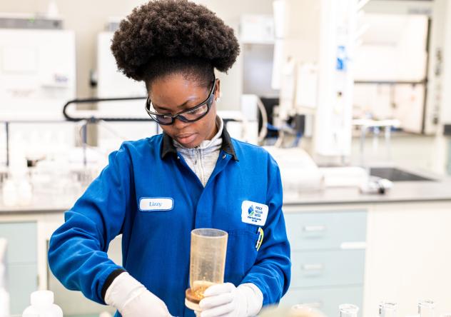 Photo: a woman in a lab coat, goggles and lab gloves working in a laboratory