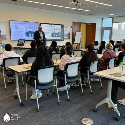A group of students listens in a classroom while a presenter gives a presentation at the front.