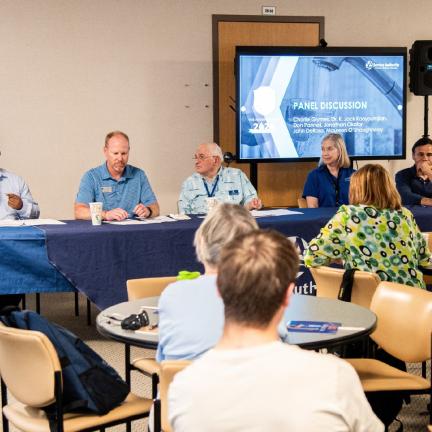 Individuals sitting at a long table presenting to a group of adults in the crowd