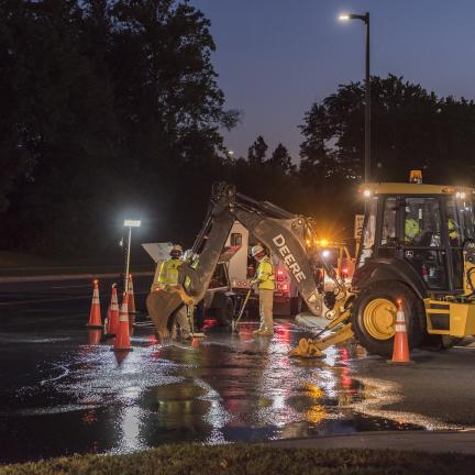 A group of utility workers stands near a backhoe at night. There is water on the ground from a water main break. Work lights illuminate the work area.