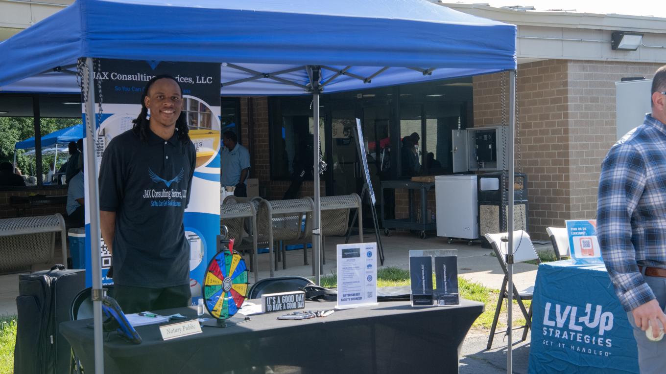 A vendor smiles behind a table under a tent at the 2026 Supplier Diversity Expo