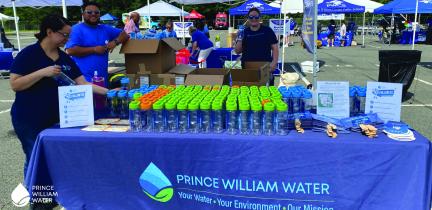 Prince William Water employees stand behind a table at an event. Reusable water bottles line the table.