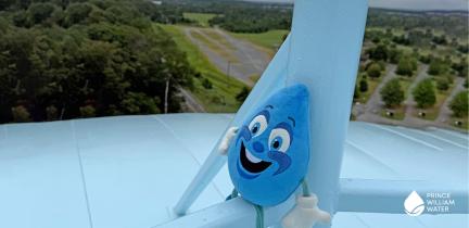 A plush water drop sitting atop a water tower with a landscape sprawled in the background