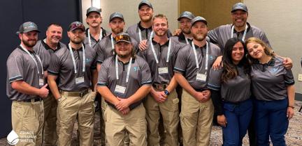 A group of individuals in matching gray polos, khaki pants and baseball hats poses in front of the camera.