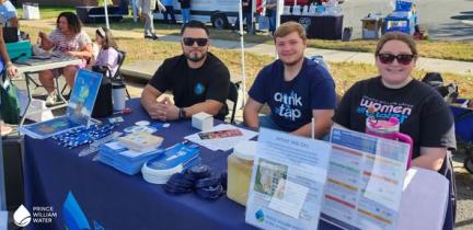 Individuals sit at a display table underneath a tent at an outdoor event