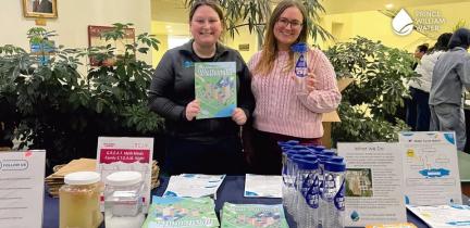 Two individuals stand behind a table of giveaways. One woman is holding a coloring book.