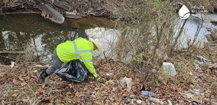 An individual in a safety jacket stooped over with a trash bag removing trash from next to a stream.