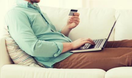 Photo: a man sitting on a couch with a laptop on his lap and credit card in hand.