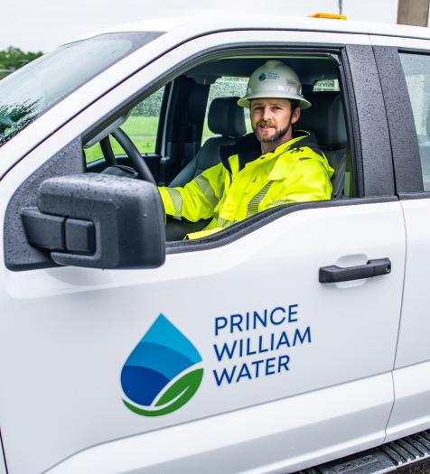 Photo: a man wearing safety gear in a white pickup truck. The truck's door has a green and blue logo with a water drop and says "Prince William Water"