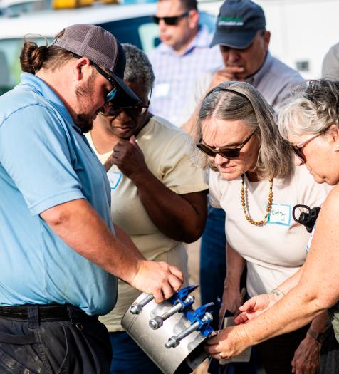 Photo: a group of people crowd around an employee showing off a piece of maintenance equipment