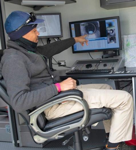 An employee sits in a chair at a computer and points to a computer screen with an image of the inside of a sewer line on screen